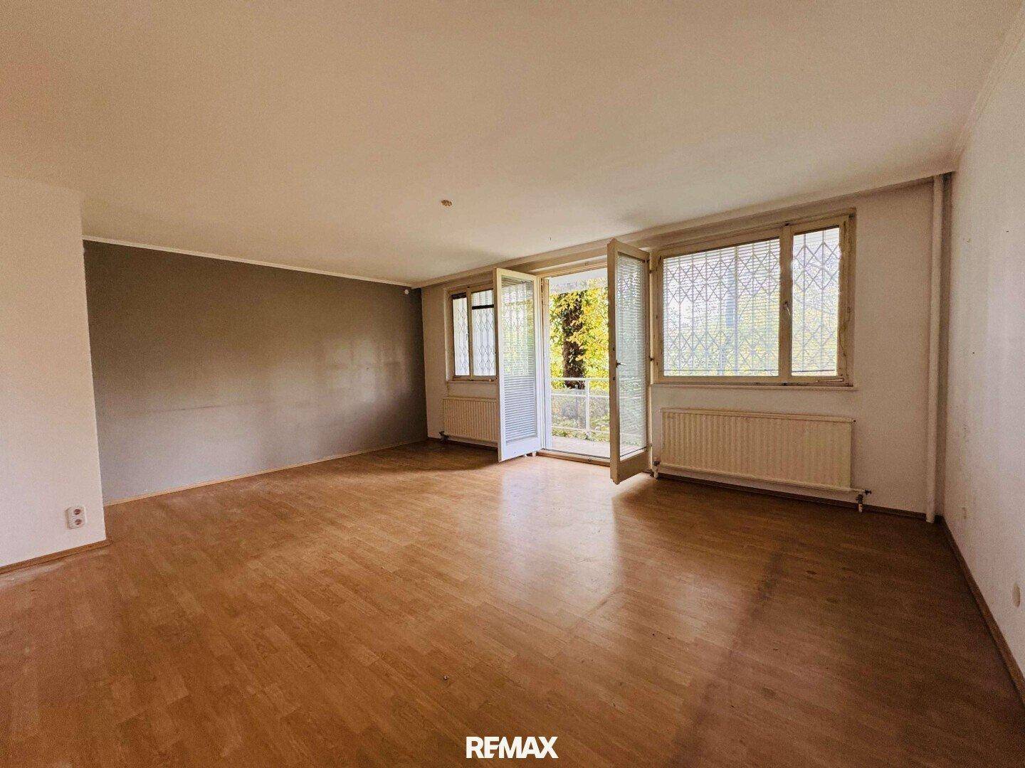 Helles, leeres Wohnzimmer mit Parkettboden und Zugang zu einem Balkon.