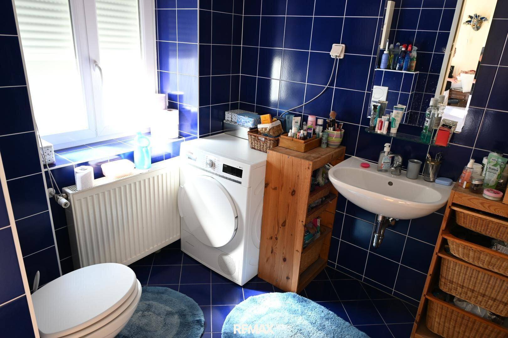 Badezimmer mit blauen Fliesen, WC, Waschbecken und Waschmaschine, hell durch ein großes Fenster.