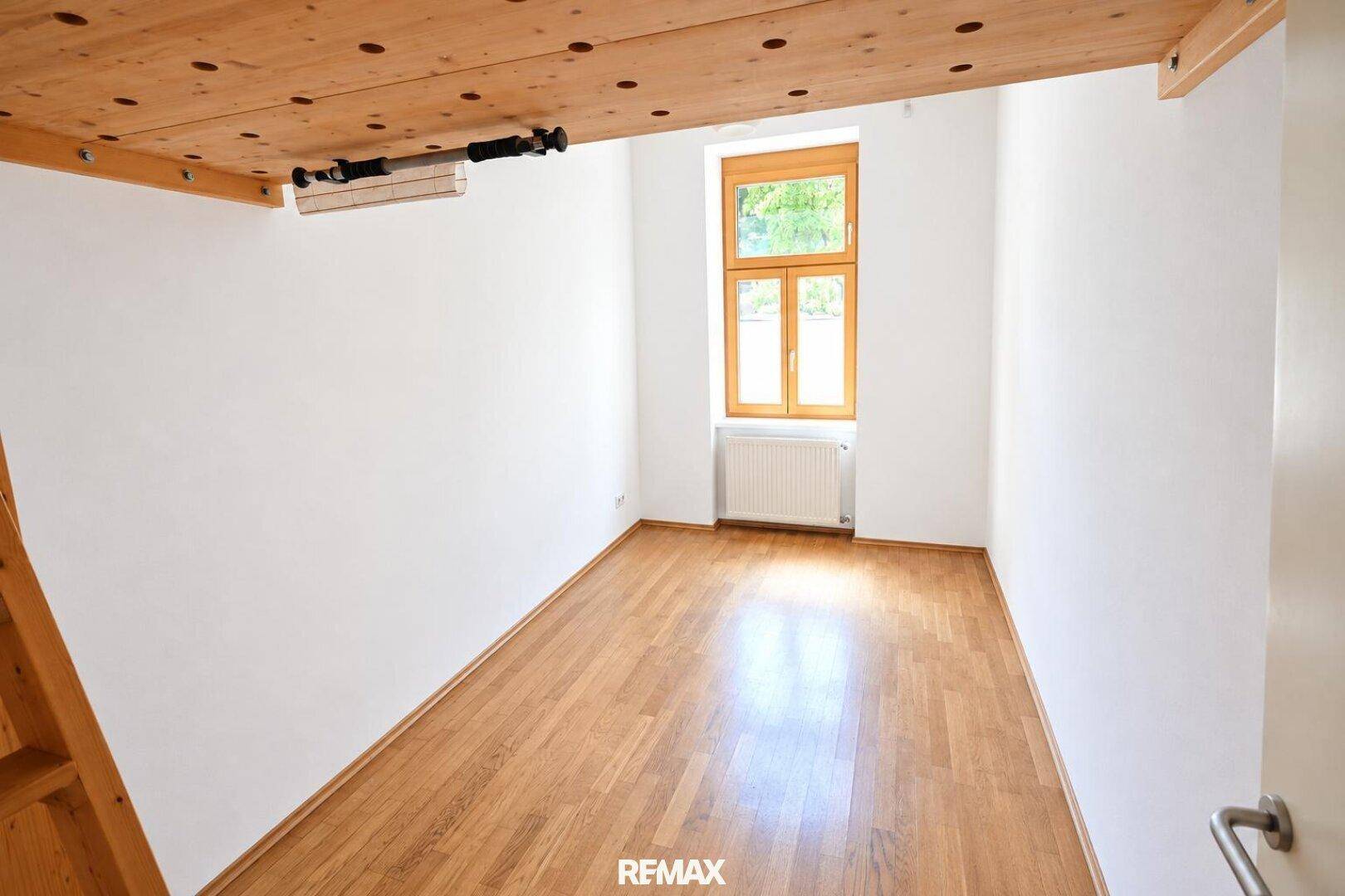 Lichtdurchflutetes Schlafzimmer mit Holzboden und großem Fenster, ideal für ein Kinder- oder Gästezimmer.
