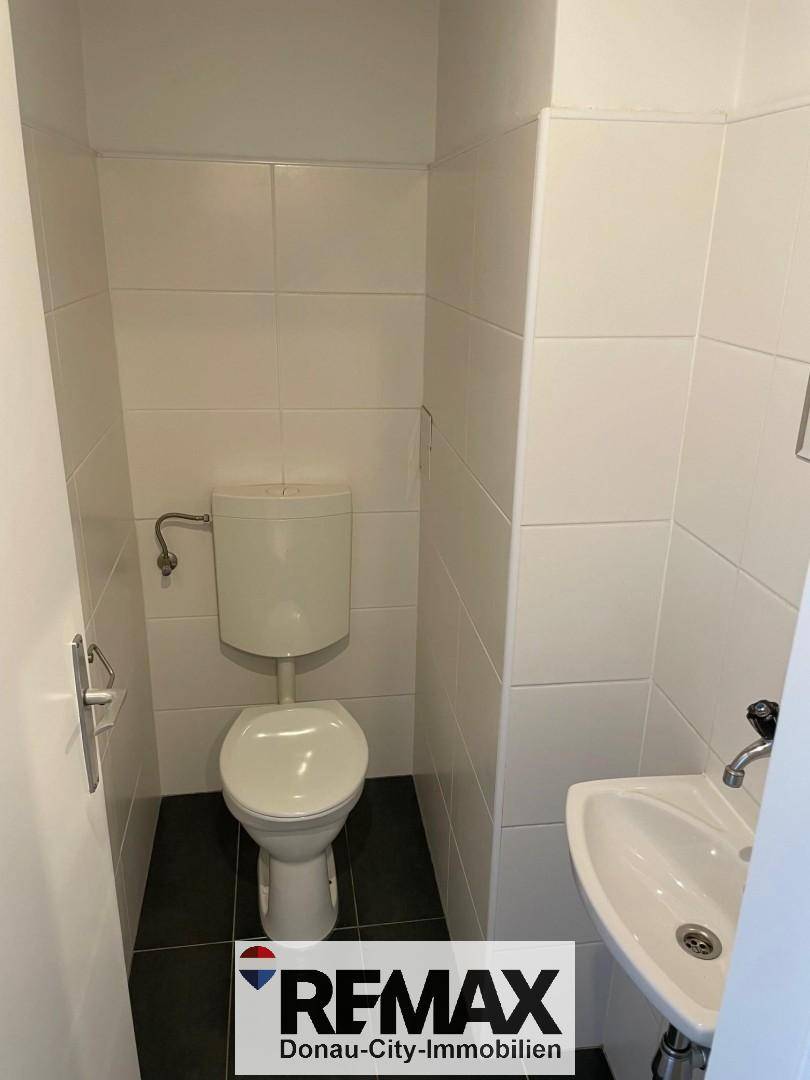 Separates WC mit Toilette, kleinem Waschbecken und weißen Fliesen an den Wänden.