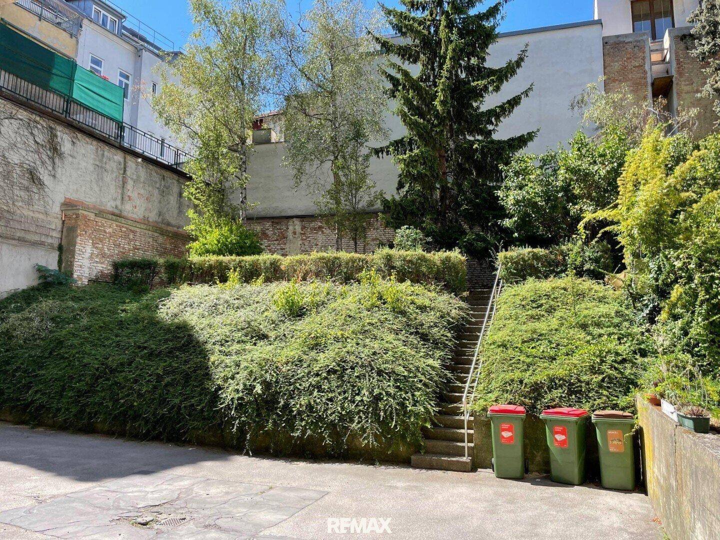 Hinterhofgarten mit Treppe, üppiger Bepflanzung und Mülltonnen in einem städtischen Umfeld.