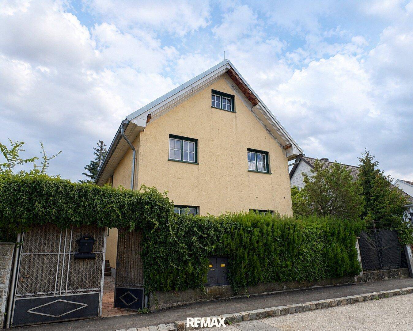 Gelbe Hausfassade mit grüner Hecke und Einfahrtstor unter bewölktem Himmel.