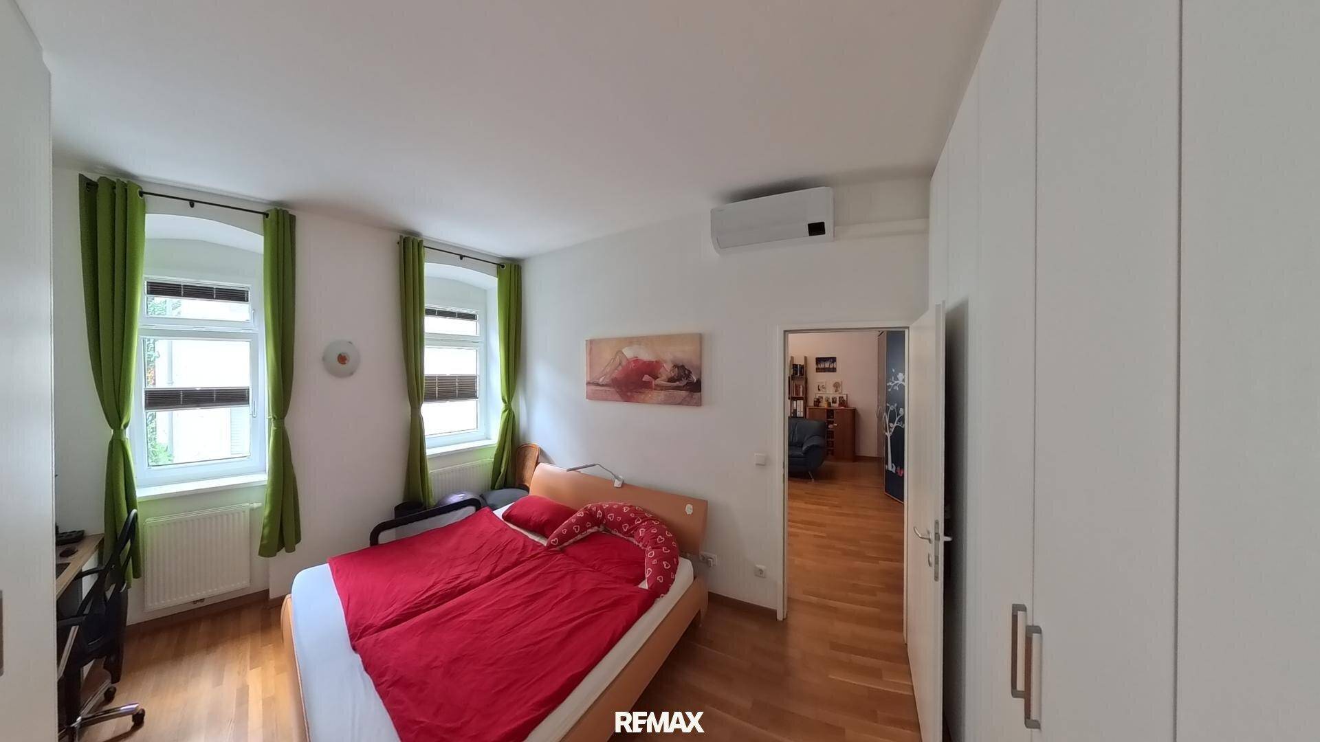 Schlafzimmer mit rotem Bett, Holzboden, grünen Vorhängen und Klimaanlage, mit Blick in einen Nebenraum.