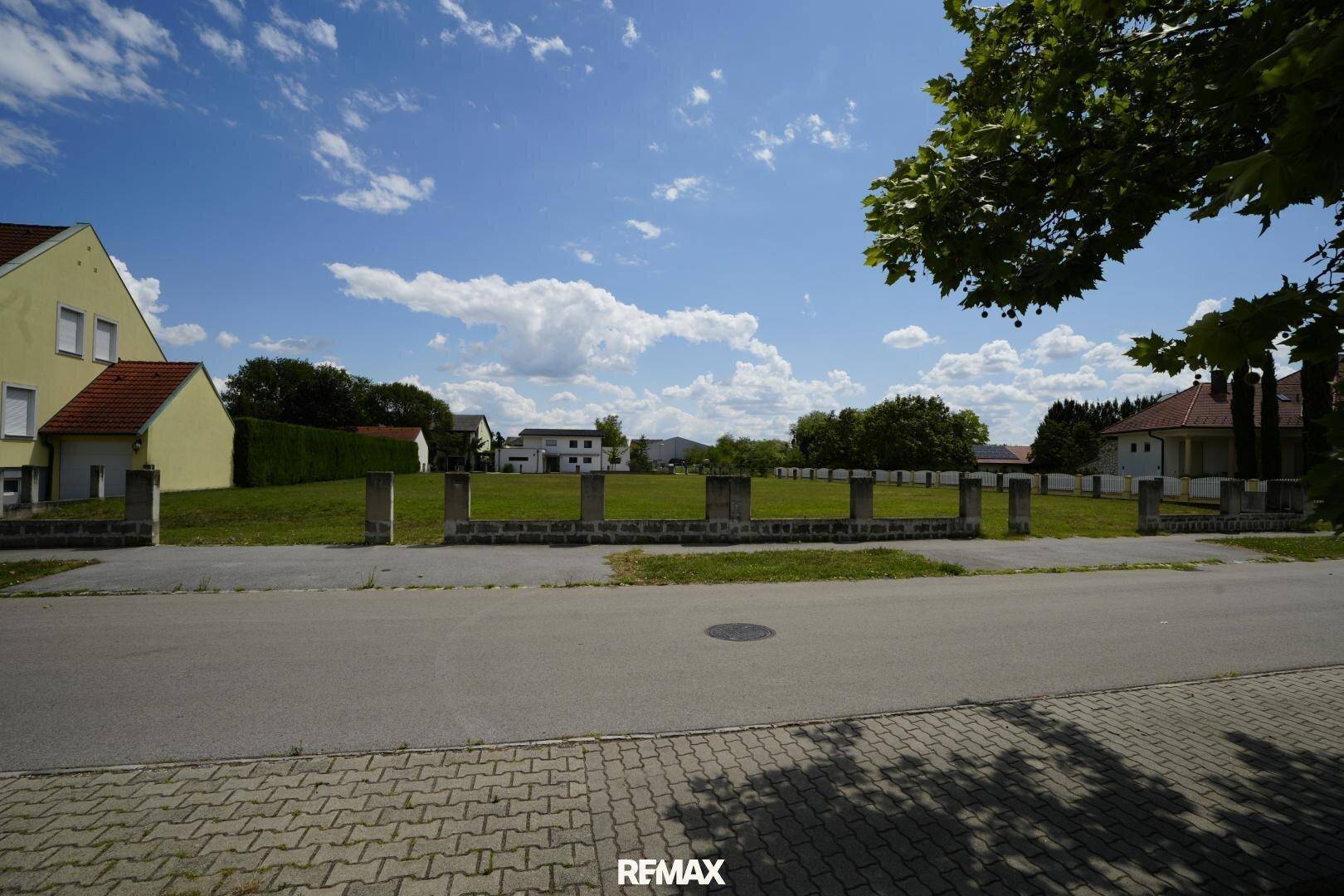 Blick von der Straße auf das grüne Grundstück mit einer niedrigen Mauer und Häusern im Hintergrund.
