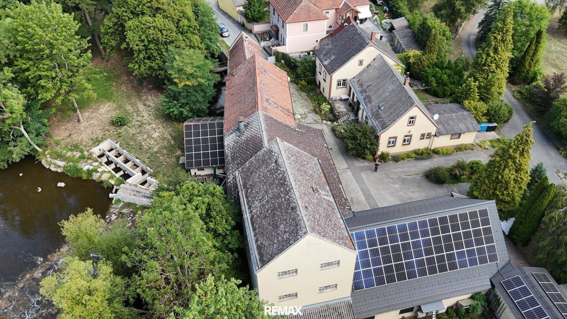 Luftaufnahme des Anwesens mit mehreren Gebäuden, Solaranlagen auf den Dächern und angrenzendem Fluss.