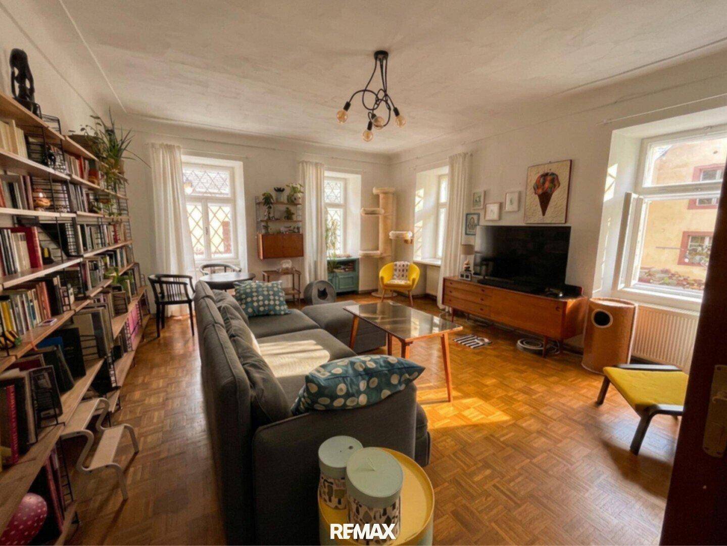 Gemütliches Wohnzimmer mit Fischgrätparkett, Bücherregal, Sofa und mehreren Fenstern für viel Tageslicht.