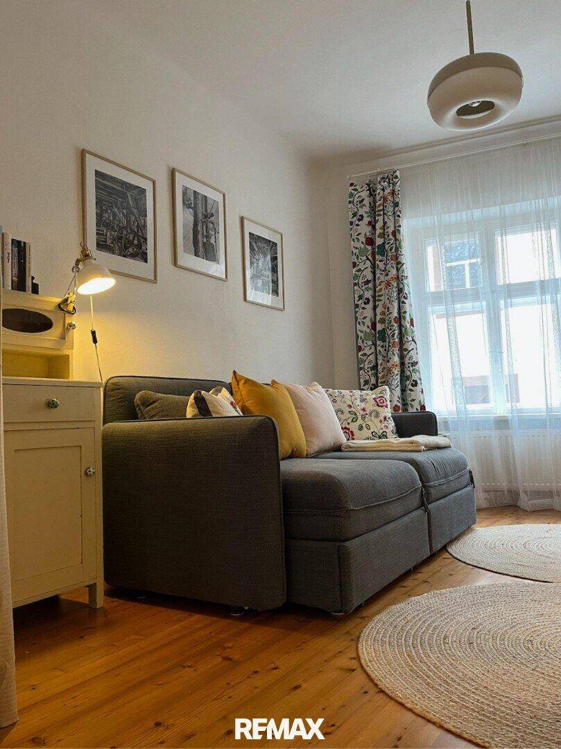 Helles Schlafzimmer mit Holzboden, einem bequemen Sofa und einem großen Fenster mit Vorhängen.