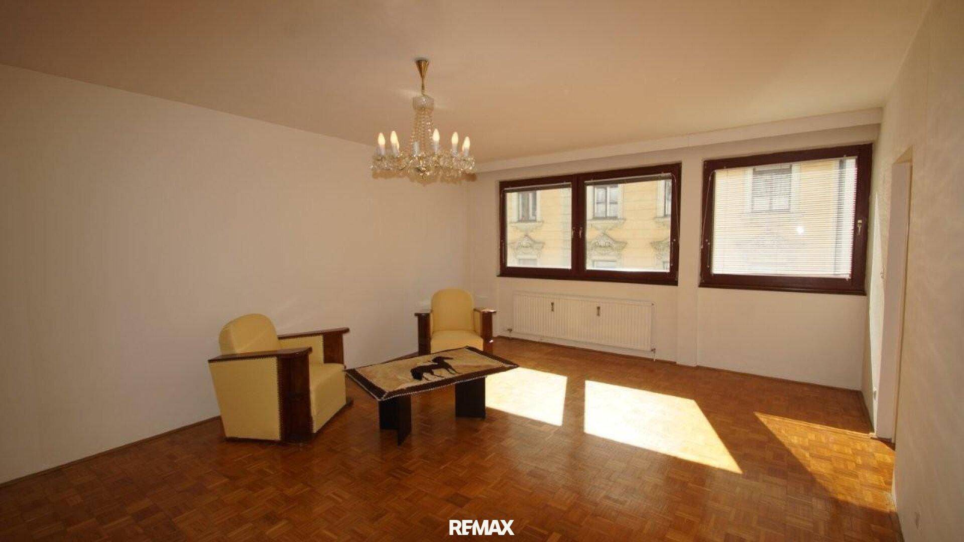 Geräumiges Wohnzimmer mit Parkettboden, Kronleuchter und zwei gelben Sesseln.