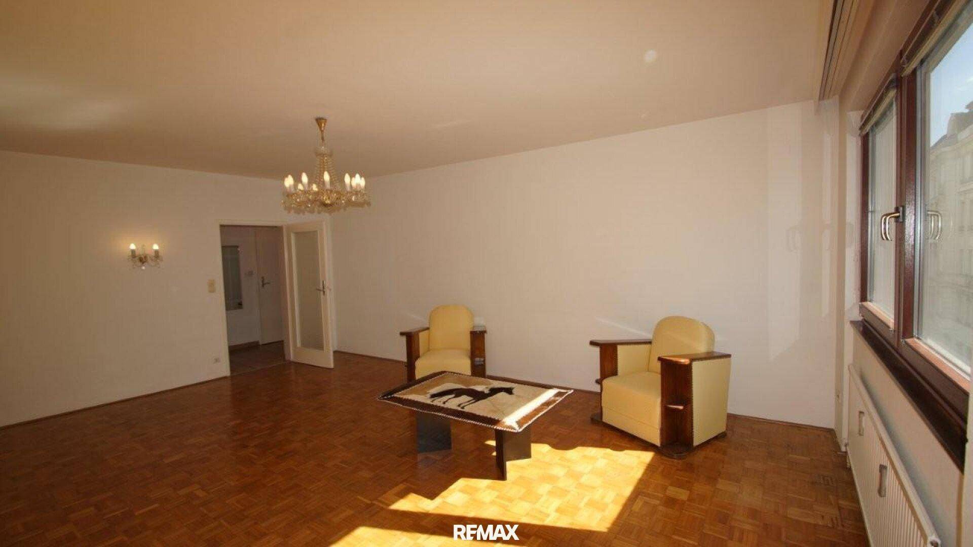 Geräumiges Wohnzimmer mit Parkettboden, Kronleuchter und zwei gelben Sesseln.