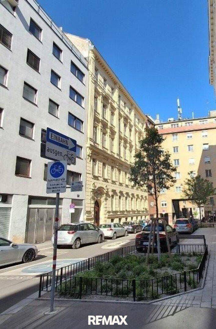Stadtansicht mit historischen und modernen Gebäuden, parkenden Autos und einem Baum.