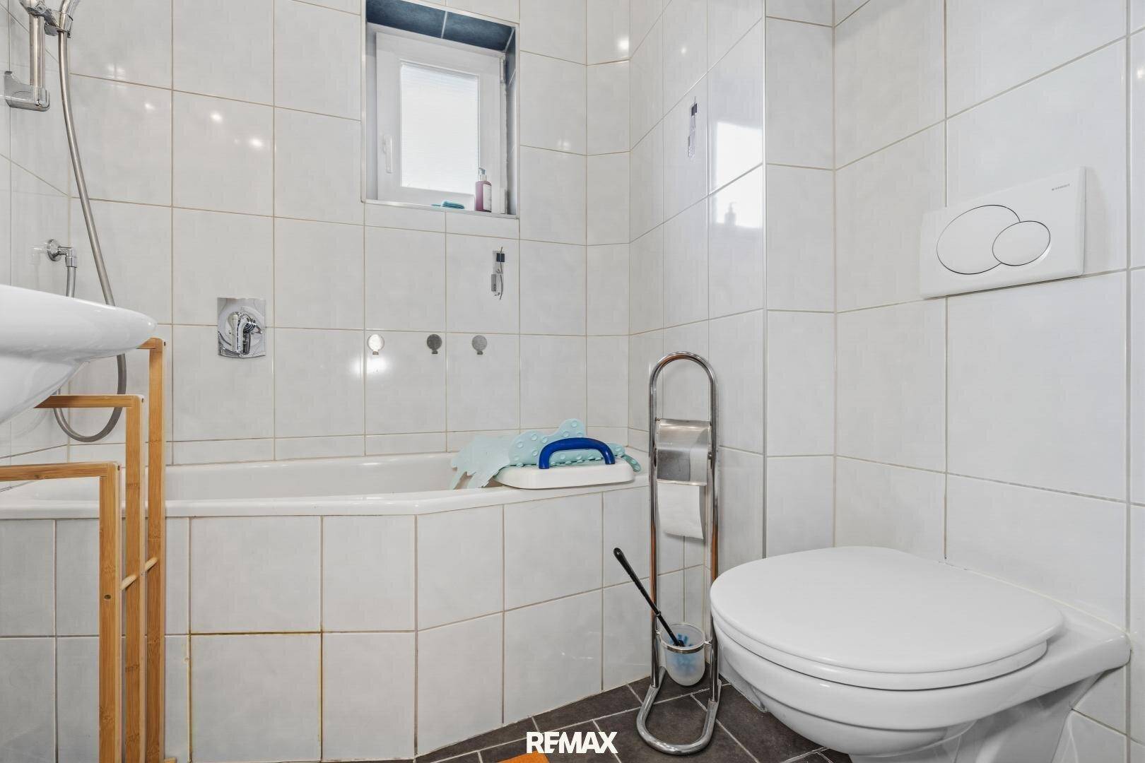 Badezimmer mit weißen Fliesen, Toilette und einer Badewanne mit Duschfunktion und Fenster.