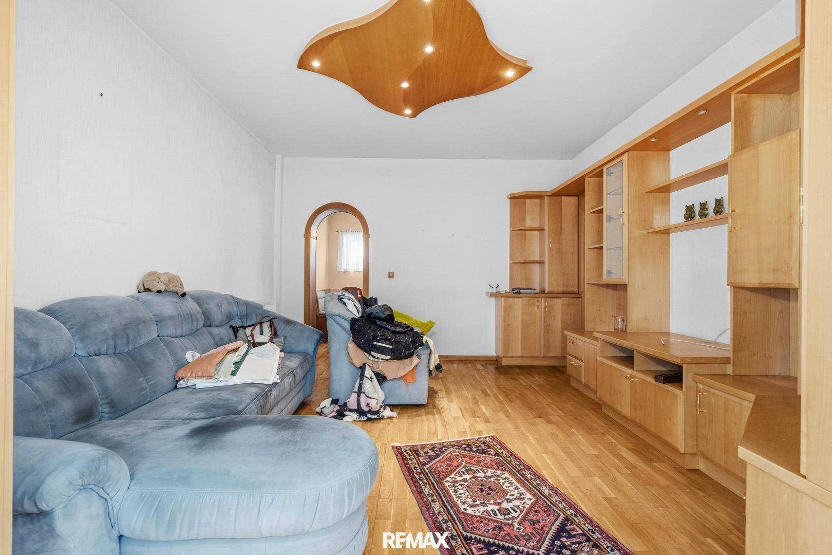 Wohnzimmer mit blauem Sofa, Holz-Wohnwand und Parkettboden, beleuchtet von einer dekorativen Deckenleuchte.