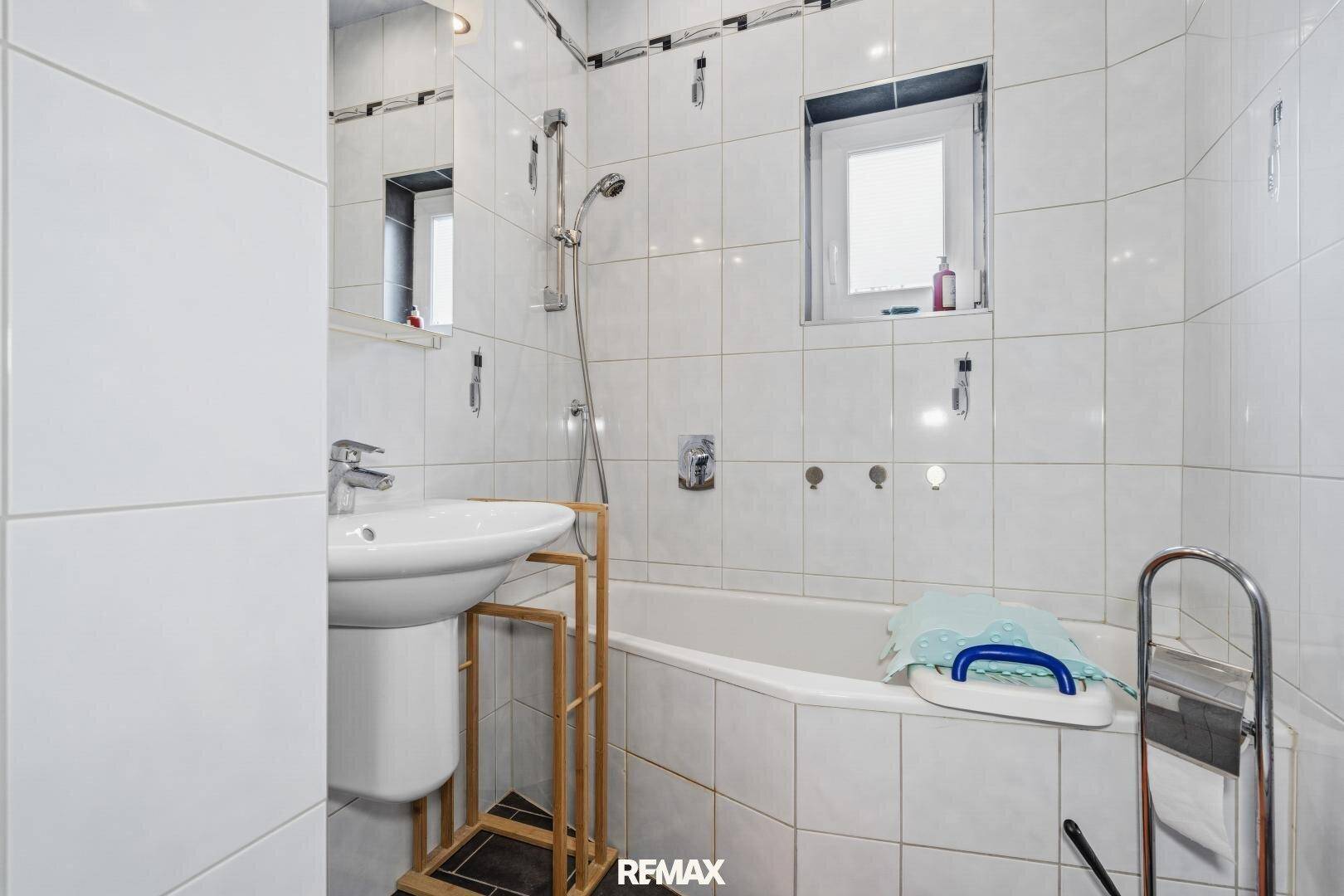 Badezimmer mit weißen Fliesen, Waschbecken und einer Badewanne mit Duschfunktion.