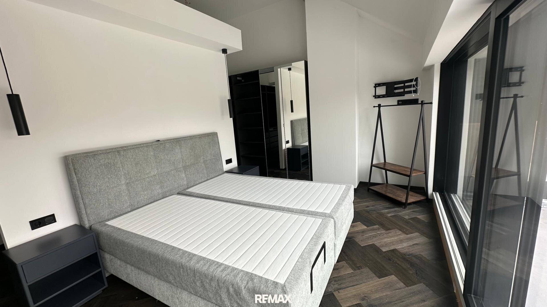 Helles Schlafzimmer mit großem Bett, dunklem Boden und bodentiefen Fenstern.