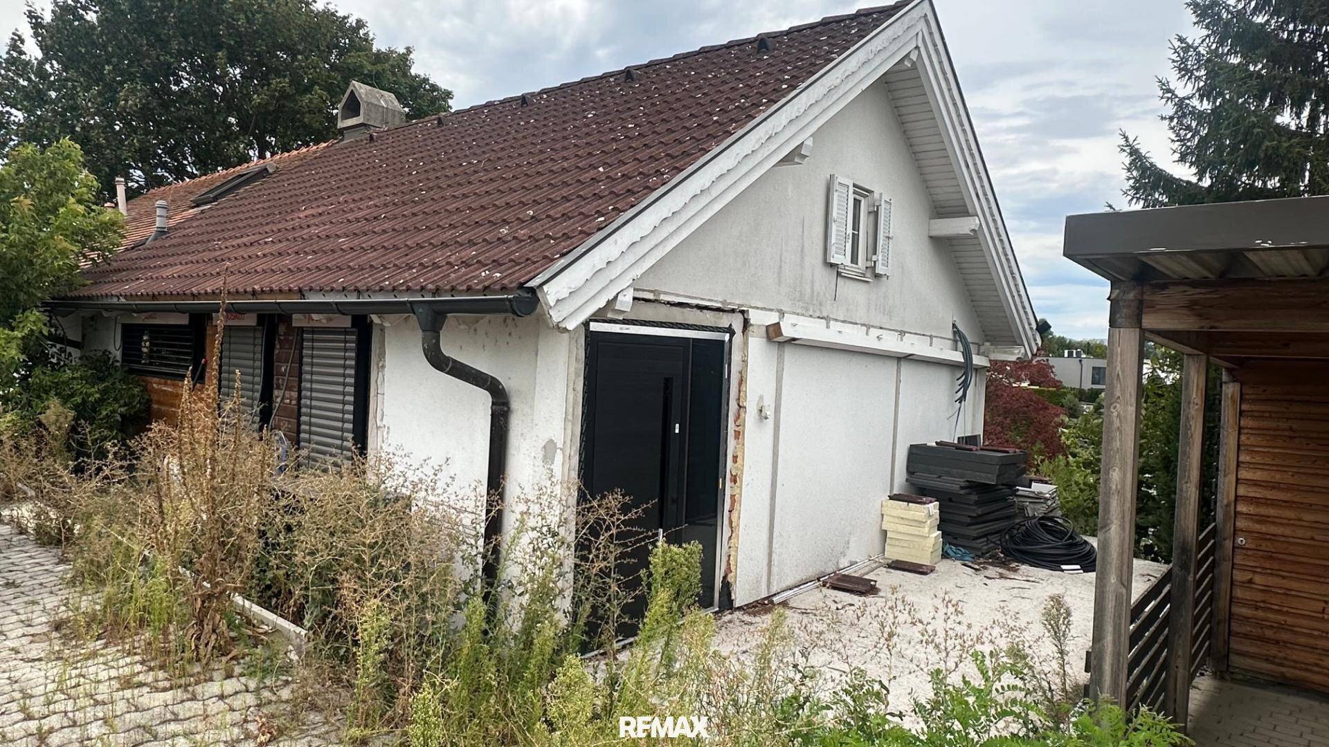 Seitenansicht eines Hauses mit Ziegeldach und überwuchertem Gartenbereich, der Renovierungsbedarf andeutet.