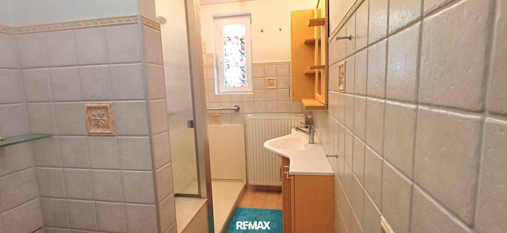 Badezimmer mit beigefarbenen Fliesen, Duschkabine, Waschbecken und Fenster.