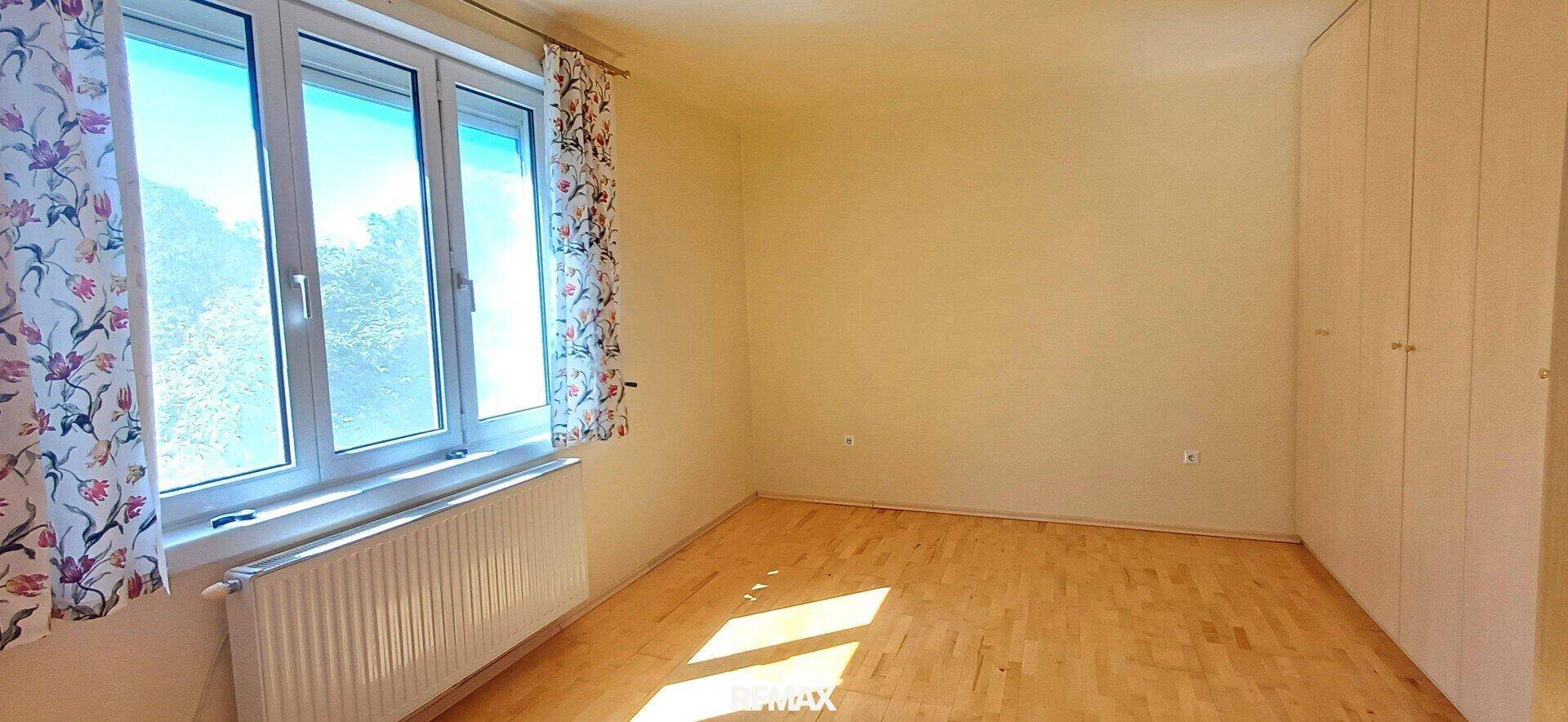 Schlafzimmer mit hellem Parkettboden, großen Fenstern und floralen Vorhängen.