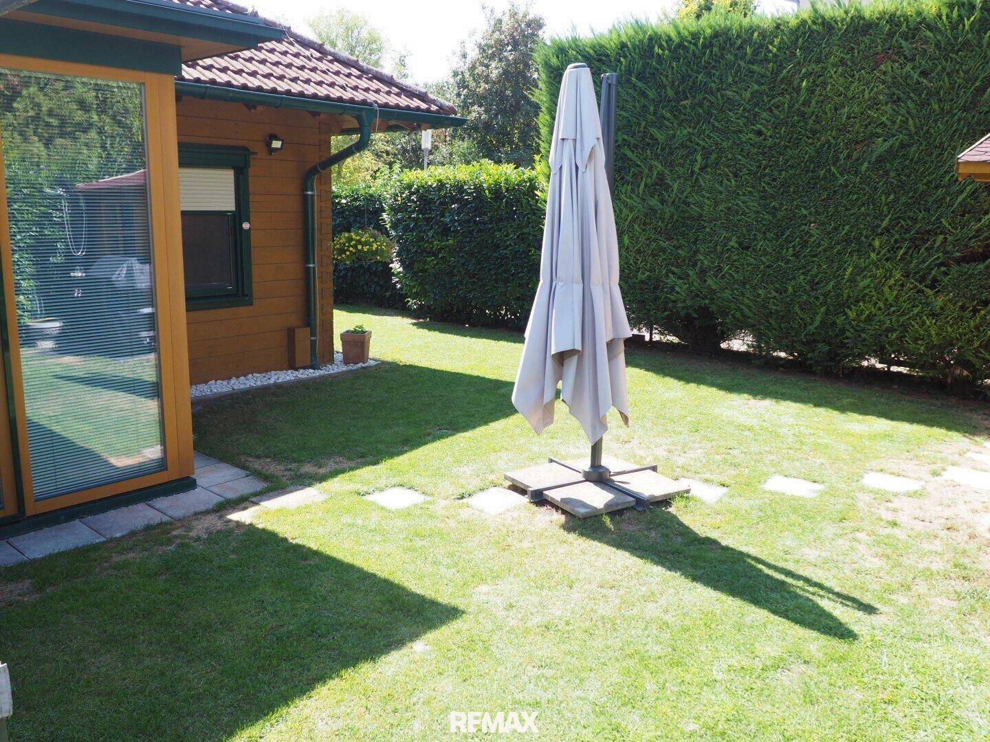 Seitlicher Gartenbereich mit Rasen, Holzhaus und großem Sonnenschirm.