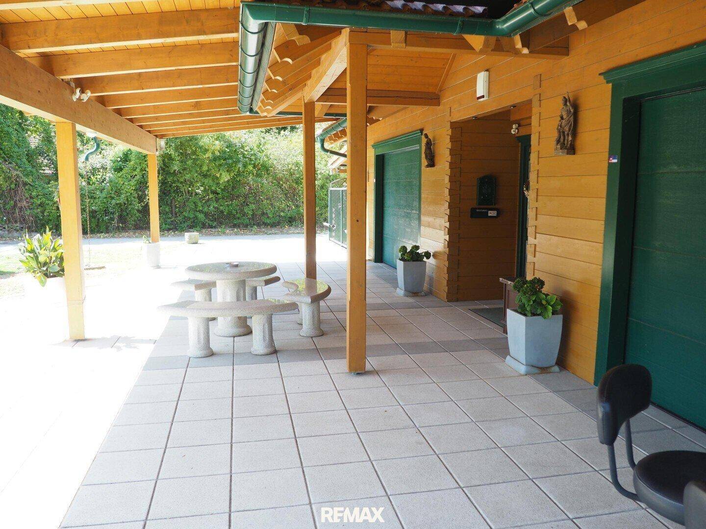 Überdachte Terrasse mit Fliesenboden und rustikaler Steinsitzgruppe vor dem Haus.