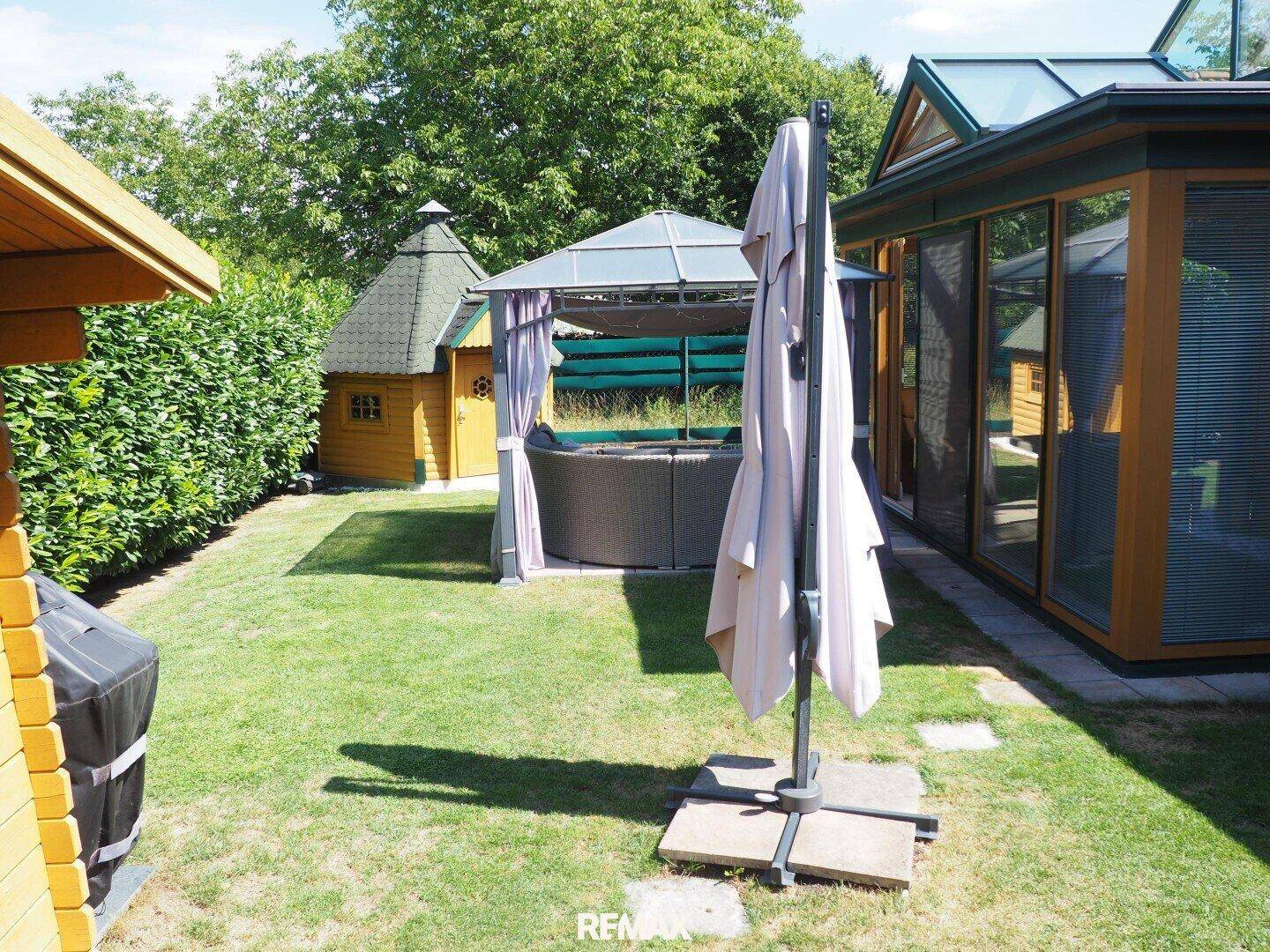 Sonniger Gartenbereich mit Wintergarten, Pavillon und großem Sonnenschirm.