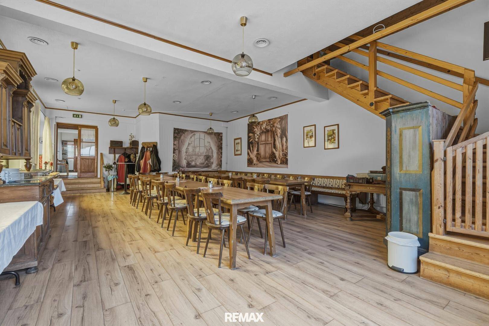 Geräumiger Speisesaal mit Holzboden, langen Tischen und einer Holztreppe.