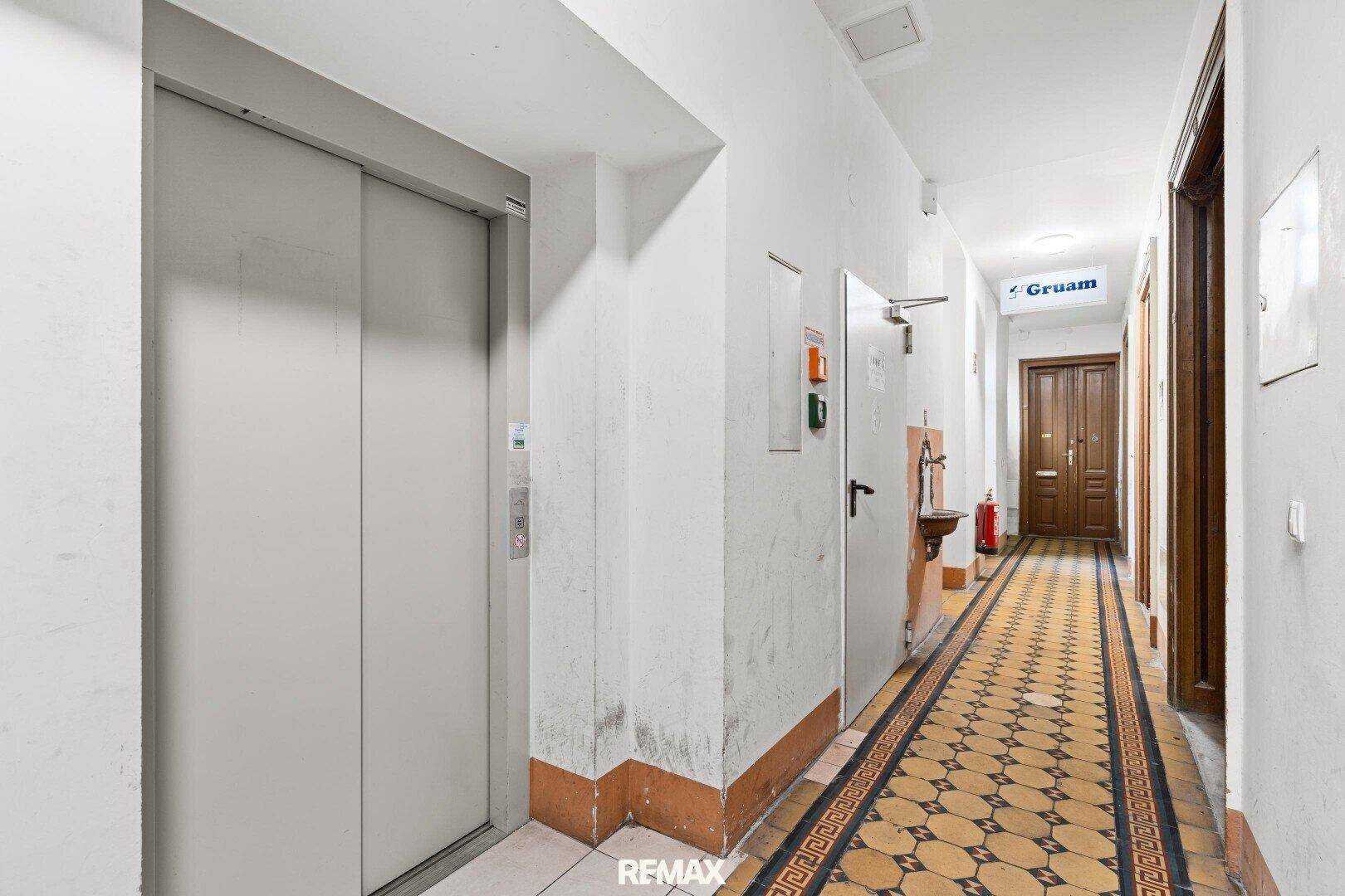 Flur im Gebäude mit Aufzug, gemustertem Fliesenboden und Holztüren.