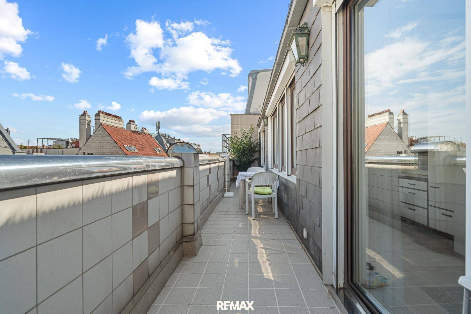 Langer, schmaler Balkon mit Fliesenboden, einer Brüstung und Blick auf umliegende Dächer.
