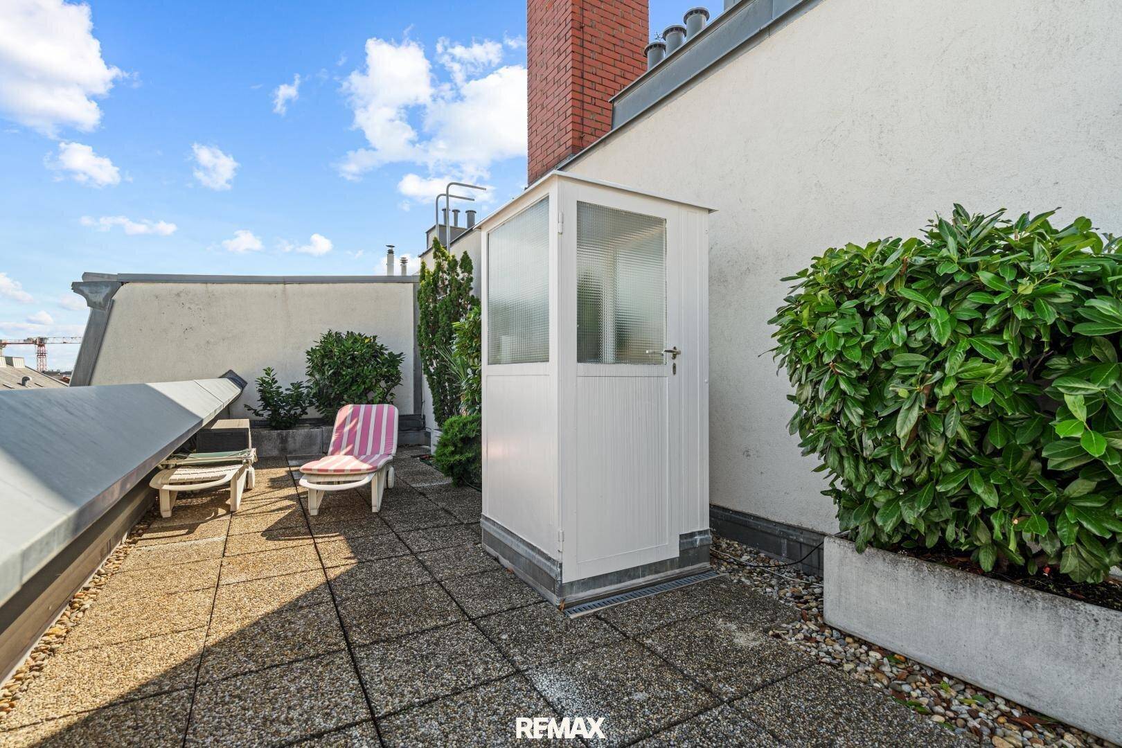 Dachterrasse mit Steinplattenboden, Liegestühlen und einer kleinen Gartenhütte unter blauem Himmel.