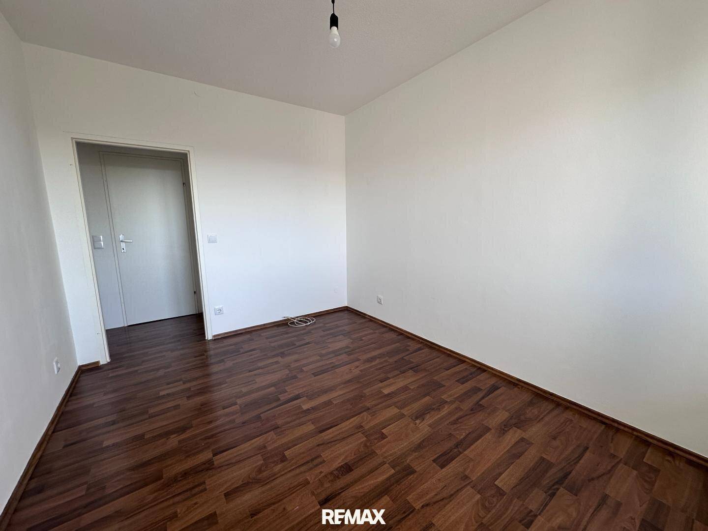 Geräumiges, leeres Zimmer mit dunklem Laminatboden und weißen Wänden.