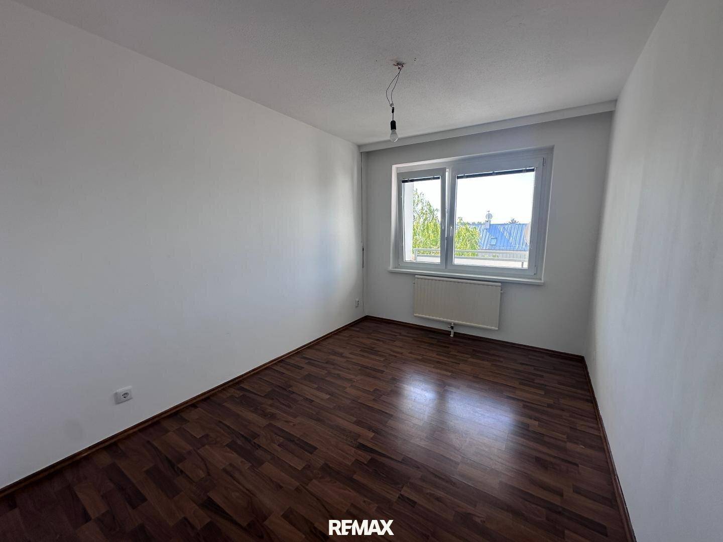 Leeres Zimmer mit dunklem Laminatboden und Fenster mit Blick ins Grüne.