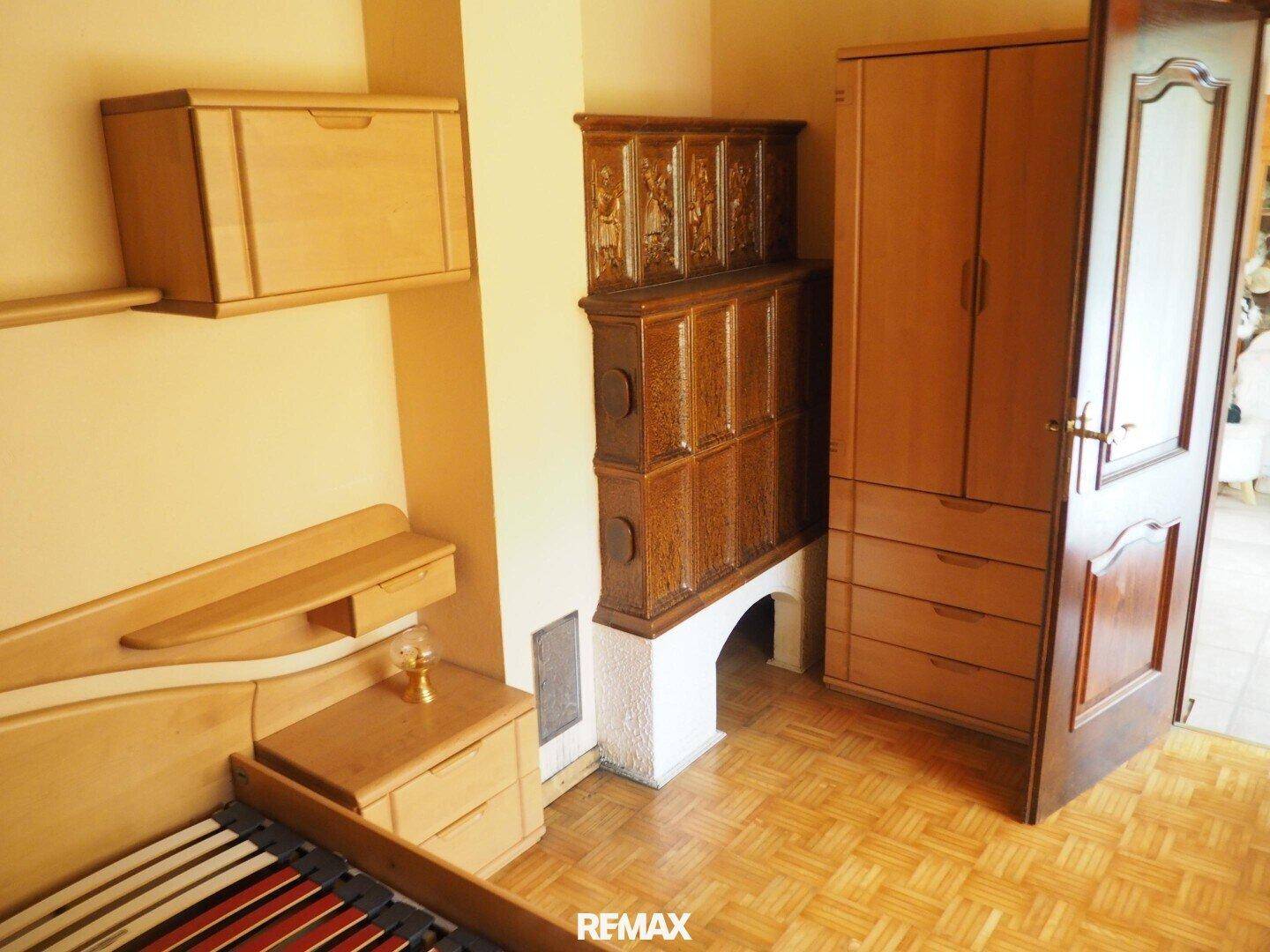 Detailansicht eines Schlafzimmers mit Holzbett, Schrank und einem Kachelofen.