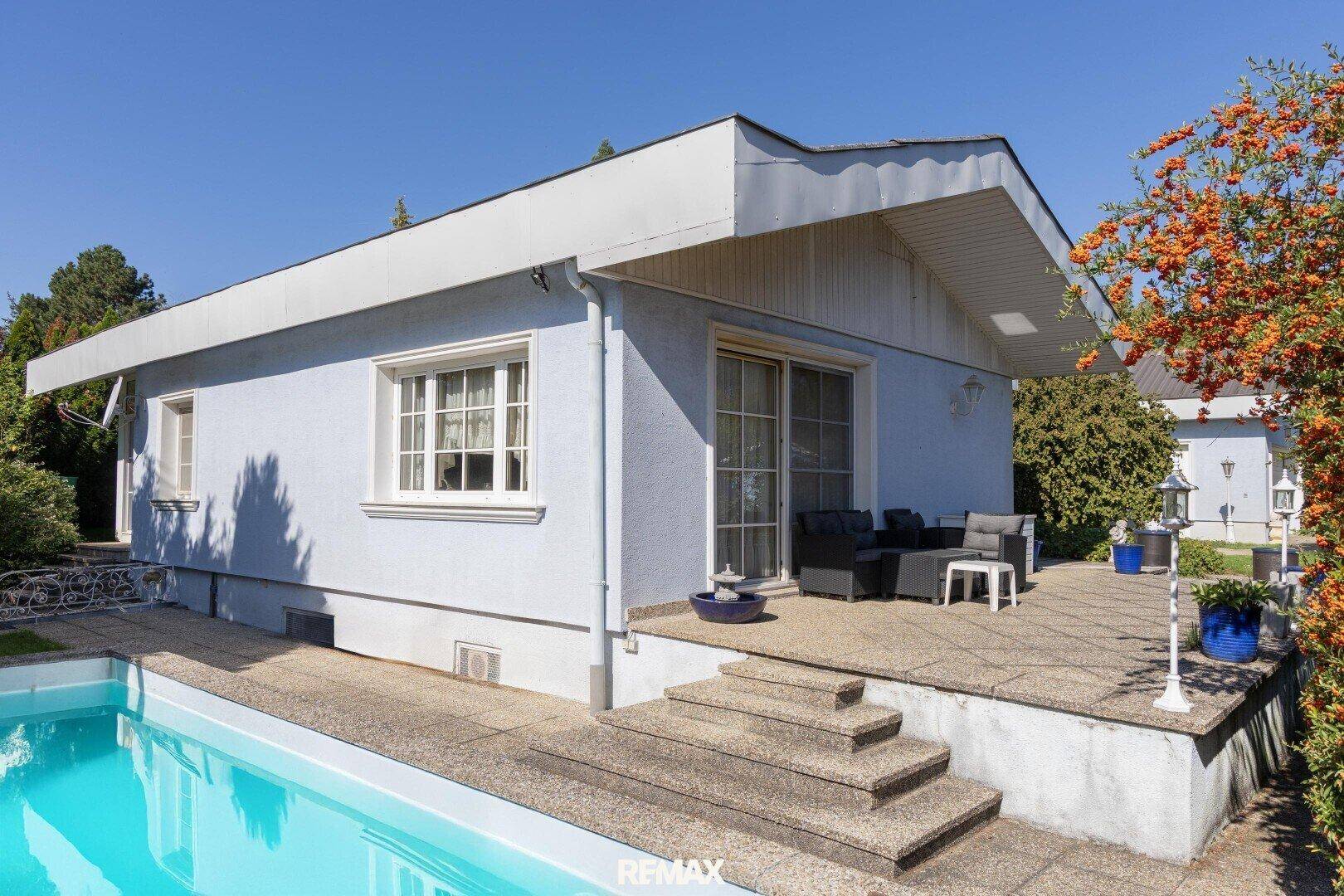 Hausfassade mit Terrasse, Pool und Sitzgelegenheiten im Freien unter blauem Himmel.