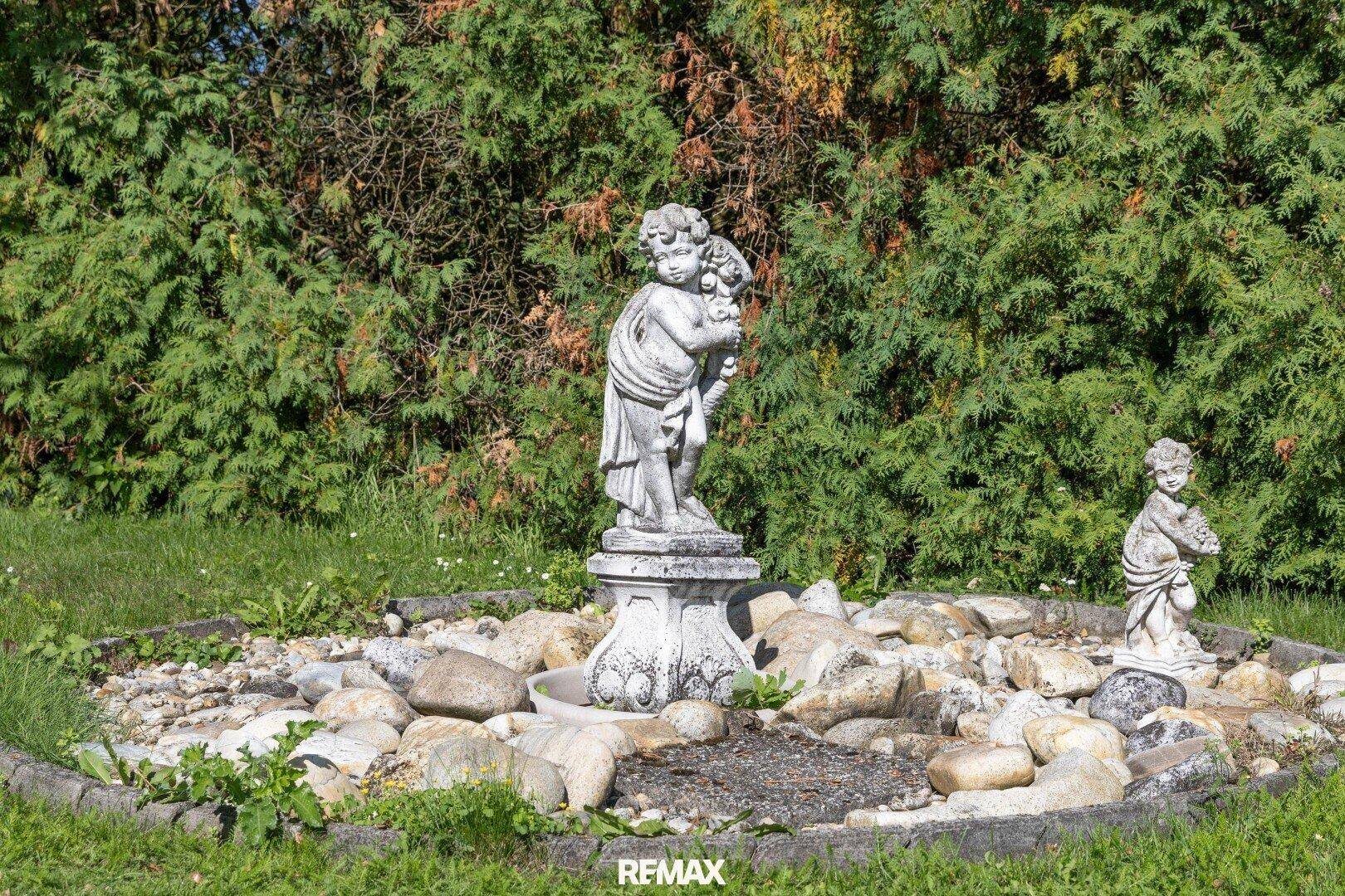 Gartenbereich mit dekorativen Steinfiguren und einem kleinen Steingarten, umgeben von Hecken.