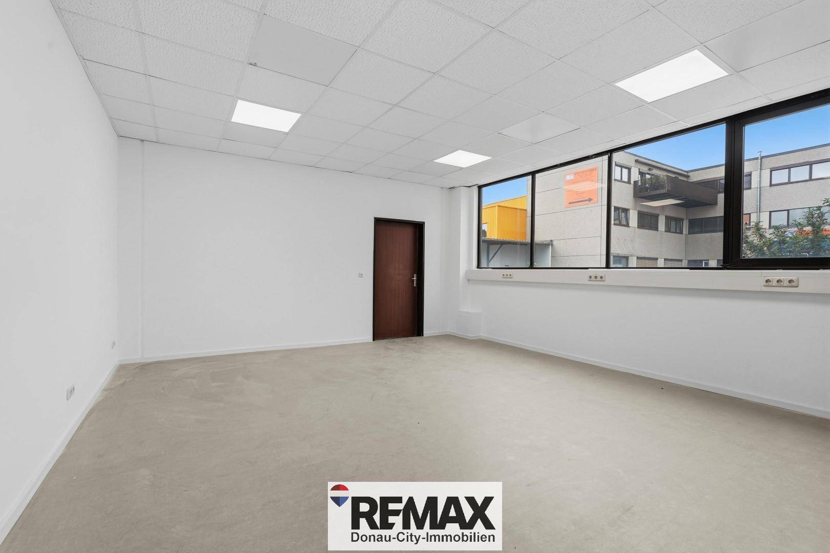 Leerer Büroraum mit weißen Wänden, abgehängter Decke und großen Fenstern mit Blick nach draußen.