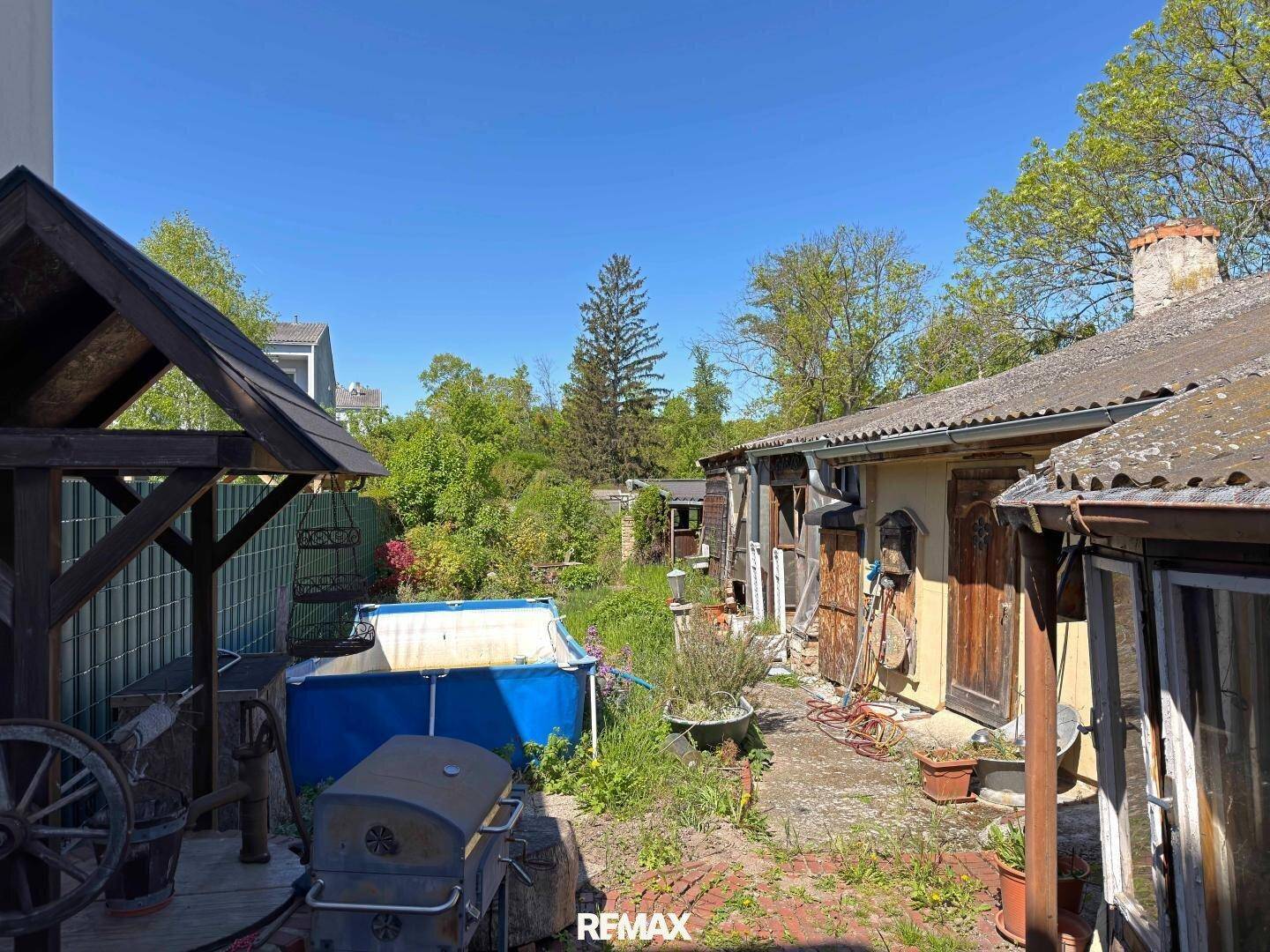 Großer Gartenbereich mit Aufstellpool, Grill und rustikalen Nebengebäuden unter blauem Himmel.