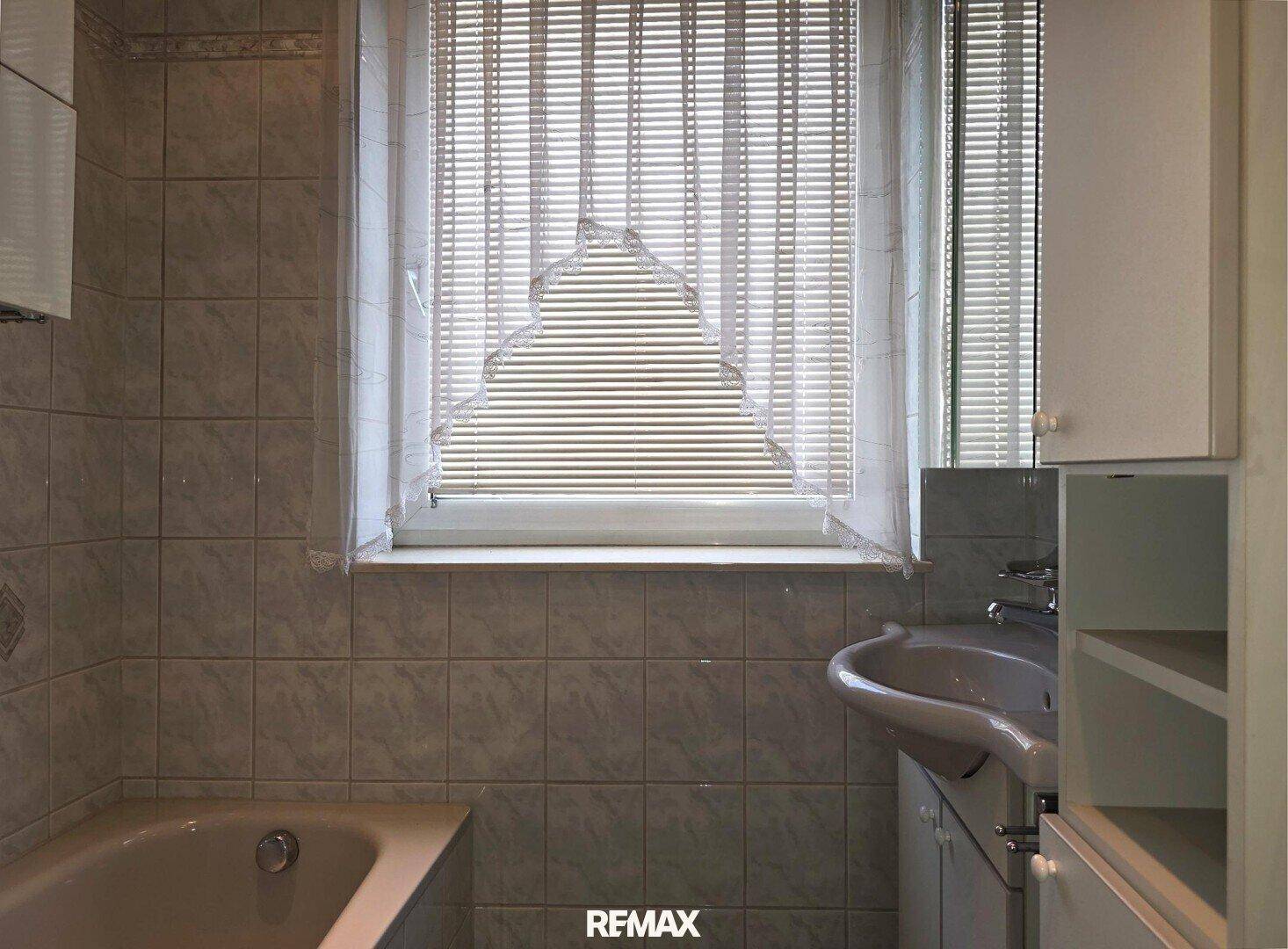 Badezimmer mit Badewanne, Waschbecken und Fenster mit Jalousien.