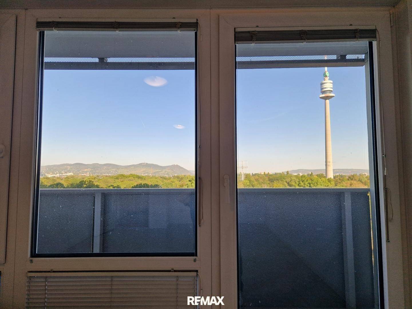 Fensterblick auf den Donauturm, grüne Landschaft und ferne Berge, mit Balkon im Vordergrund.