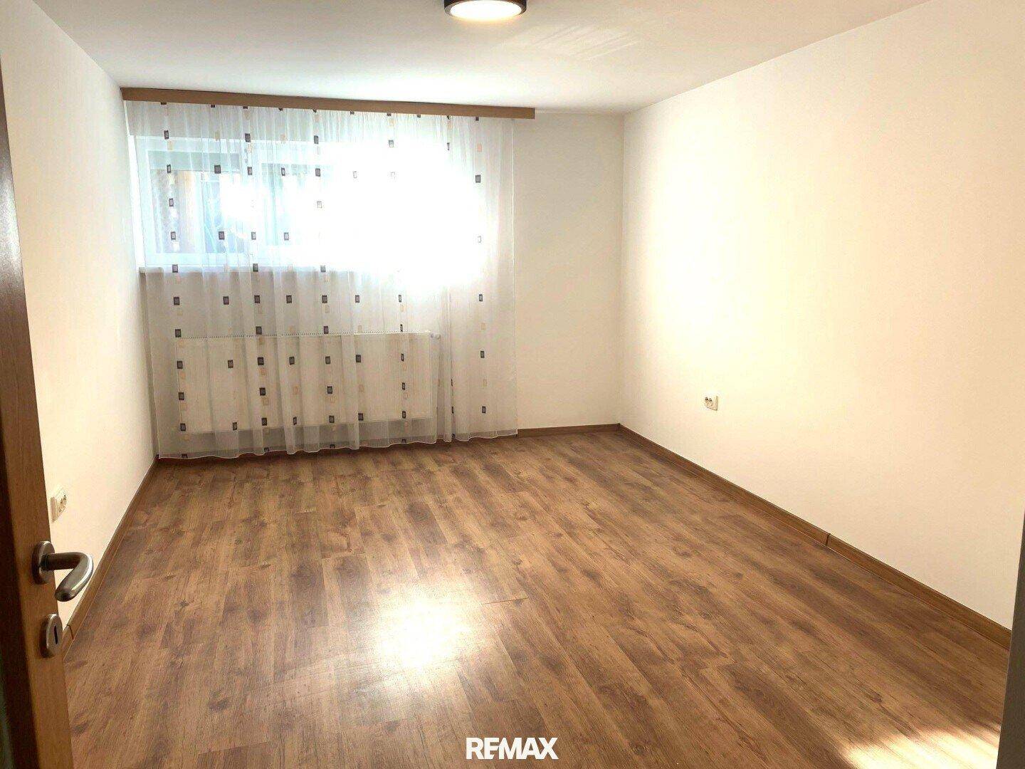 Leeres Zimmer mit Holzfußboden und einem großen Fenster, das viel Tageslicht hereinlässt.