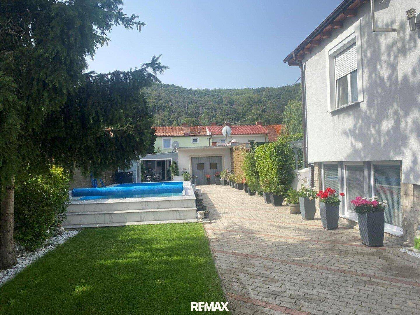 Außenbereich des Hauses mit Swimmingpool, gepflegtem Rasen und gepflasterter Auffahrt vor hügeliger Landschaft.