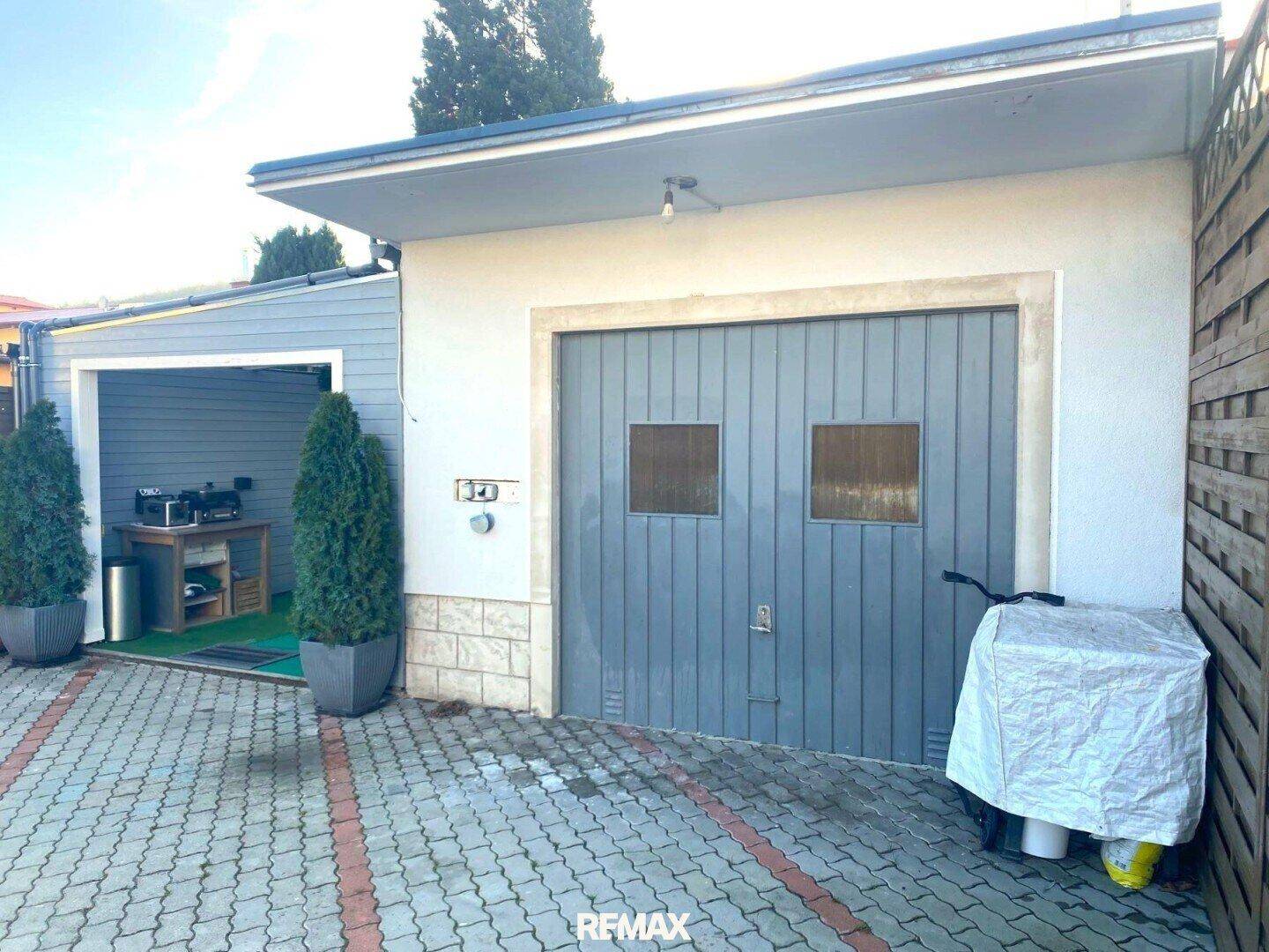 Graue Garage mit gepflasterter Einfahrt und angrenzendem Carport mit kleiner Außenküche.