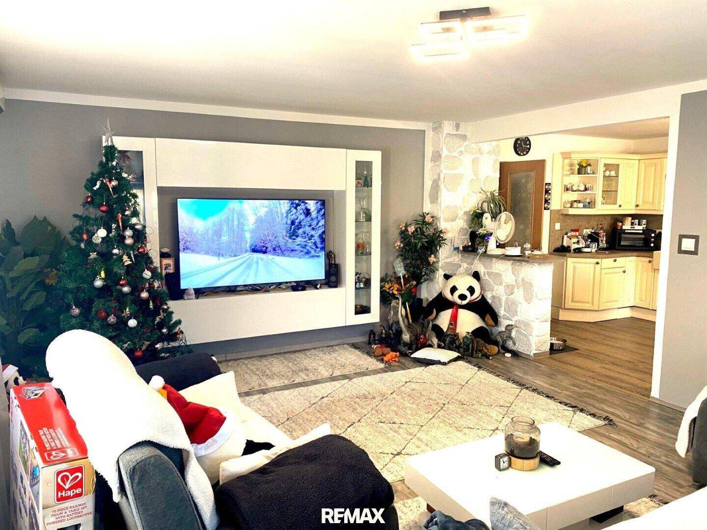 Wohnzimmer mit großem TV-Möbel, bequemer Couch und offenem Übergang zur Küche, festlich dekoriert.