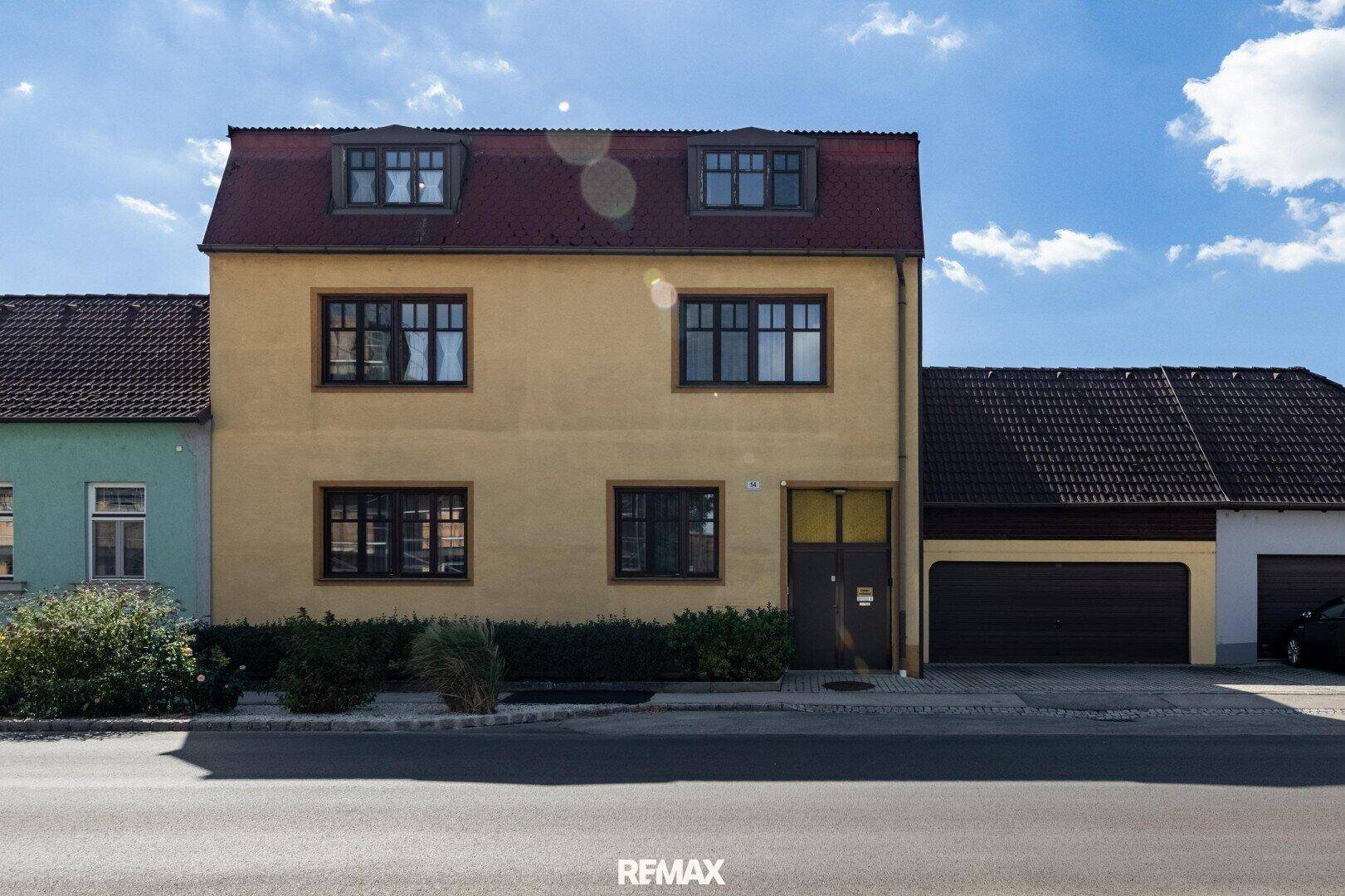 Vorderansicht eines gelben Hauses mit rotem Mansarddach, Fenstern und einer Garage.