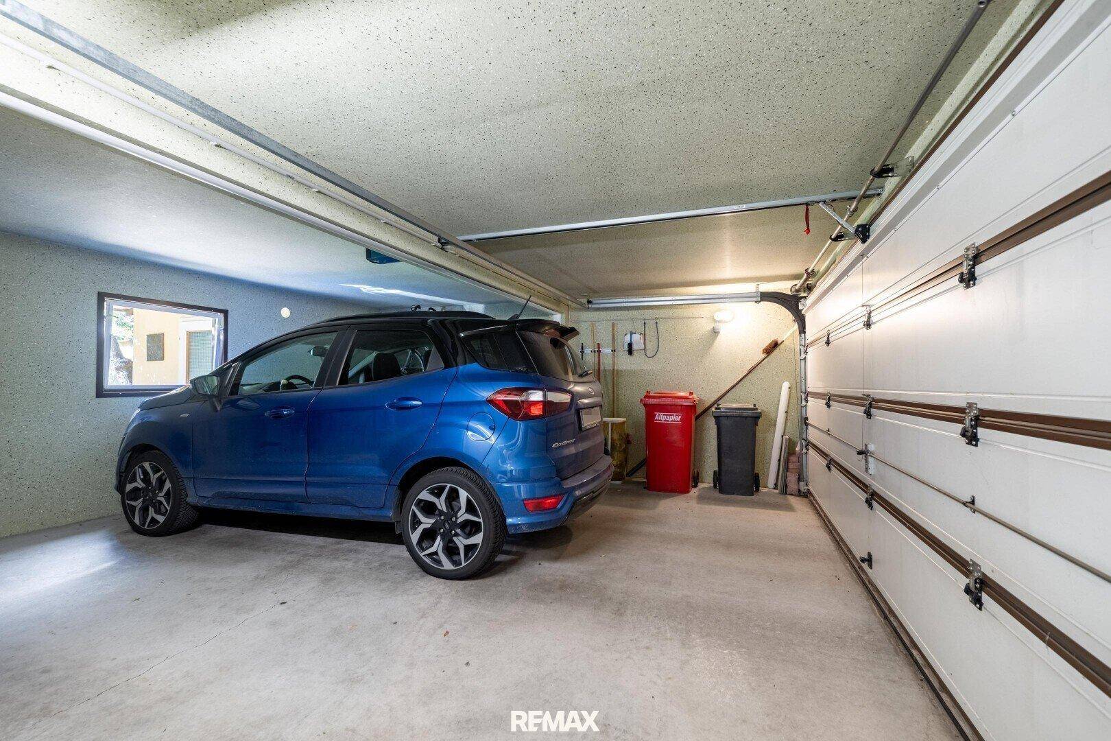 Geräumige Garage mit einem blauen Auto, weißen Garagentoren und einem Fenster.