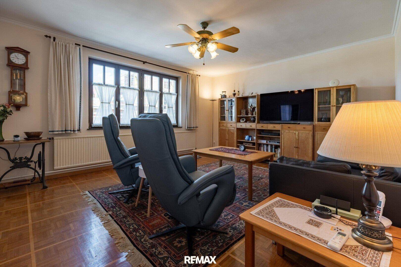 Gemütliches Wohnzimmer mit Parkettboden, Deckenventilator und großem Fenster.