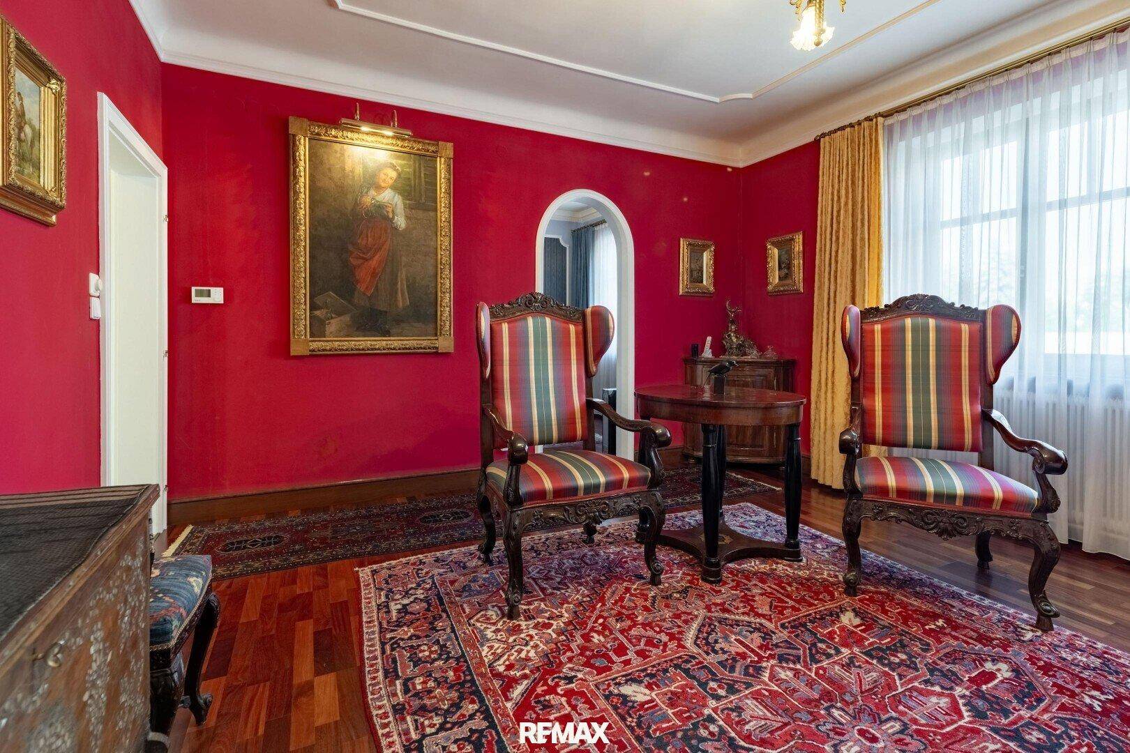 Geräumiges Wohnzimmer mit roten Wänden, antiken Möbeln und einem Durchgang zu weiteren Räumen.