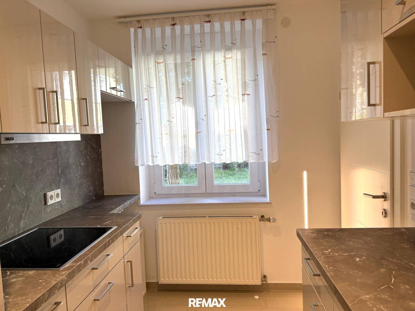 Moderne Küche mit hellen Hochglanzschränken, dunkler Arbeitsplatte und Induktionskochfeld am Fenster.