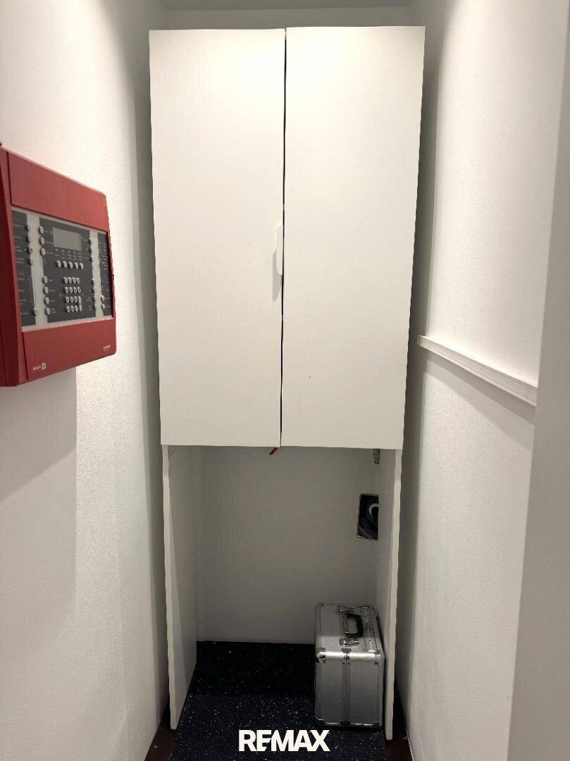 Kleiner Abstellraum mit weißen Hängeschränken und einem Brandschutzpanel an der Wand.