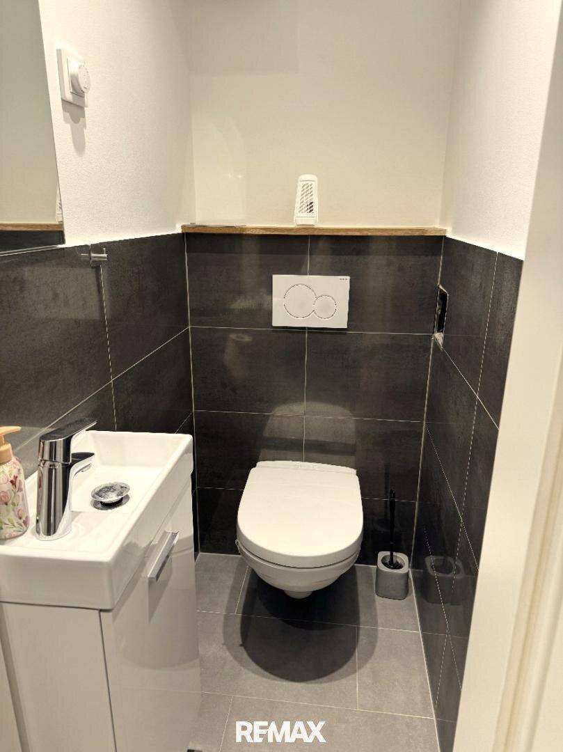 Separates WC mit wandhängender Toilette, kleinem Waschbecken und modernen dunklen Fliesen.