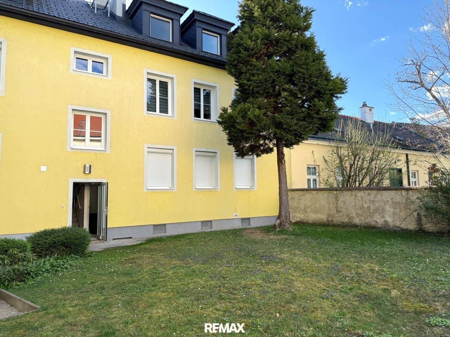 Gelbes Mehrfamilienhaus mit weißen Fenstern und einem gepflegten Vorgartenbereich mit Baum.