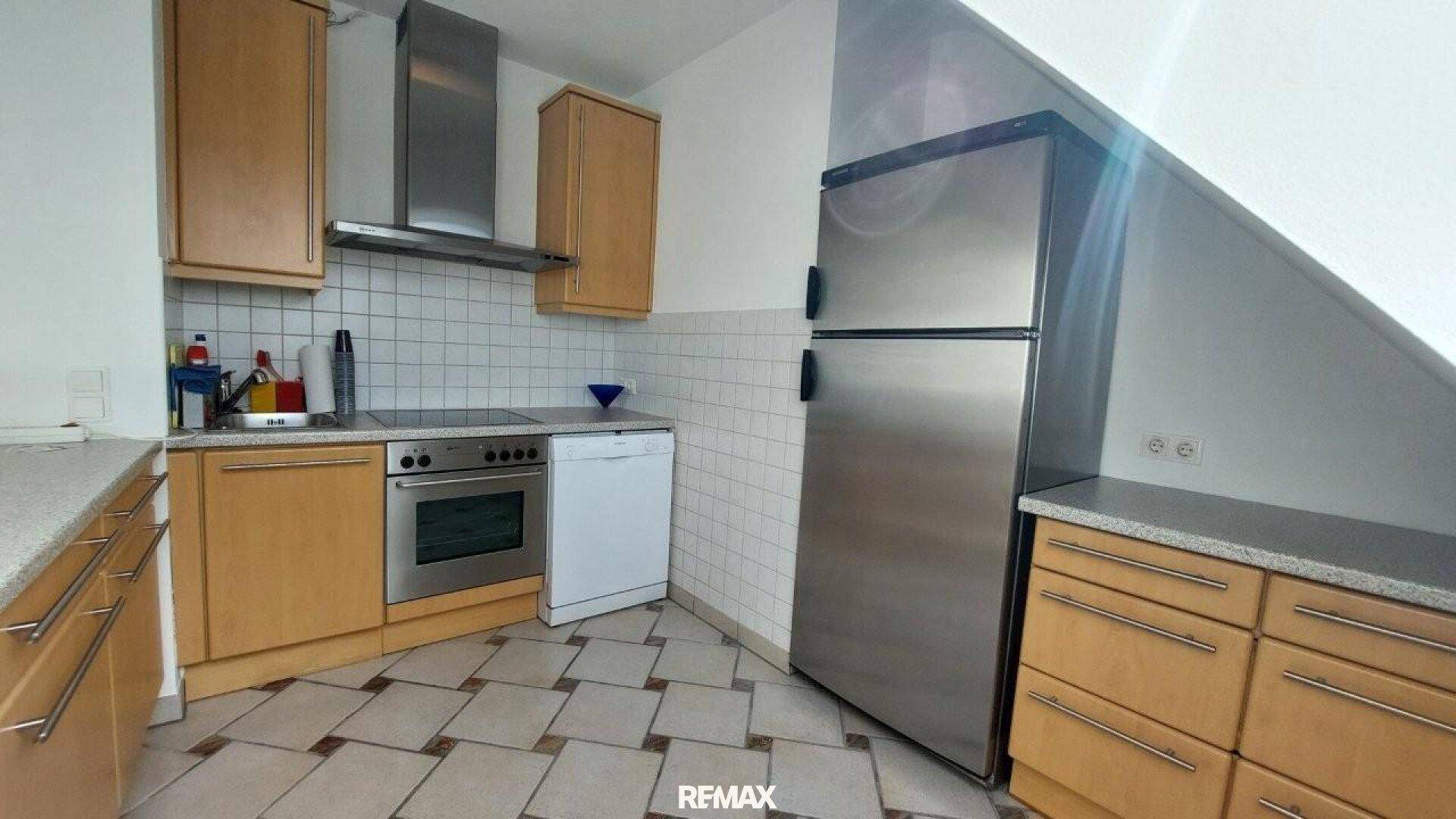 Funktionale Küche mit Holzschränken, Edelstahlgeräten und gefliestem Boden unter Dachschräge.